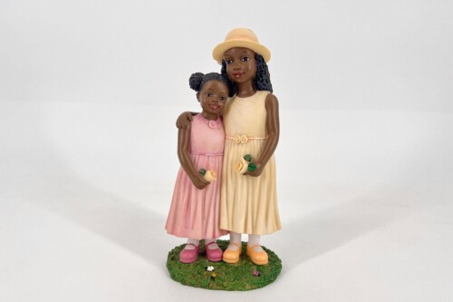 Two female figurines standing together: a taller girl in a cream-yellow dress and hat, and a shorter girl in a pink dress, each holding small flowers on a grassy base.