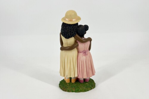 Back view of two girls figurines embracing, one in a yellow dress with a sunhat and the other in a pink dress, standing on a green base