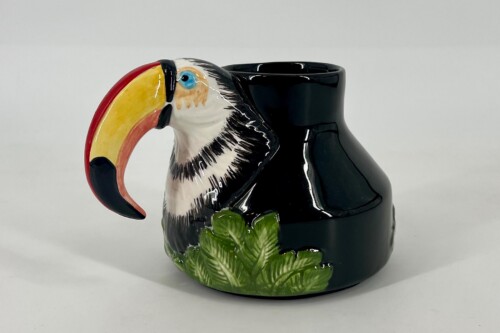 Toucan-themed ceramic mug with a large colorful beak and black body, sitting among green leaves.