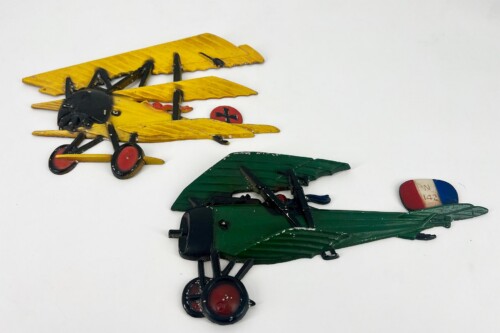 Colorful vintage toy airplanes arranged on a white surface: a yellow biplane with red wheels and a black frame, and a green plane with a red-white-blue tail fin, showing wear and chipped paint.