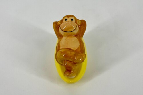Orange ceramic monkey with hands raised behind its head, sitting on a yellow oval base.