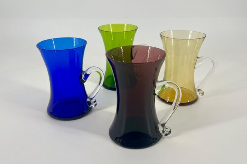 Four colored glass pitchers with clear handles arranged on a white surface: blue, green, brown, and pale yellow.