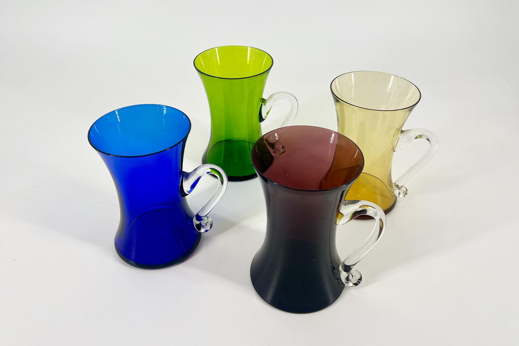 Four colored glass pitchers (blue, green, amber, and brown) with clear handles arranged on a white surface.