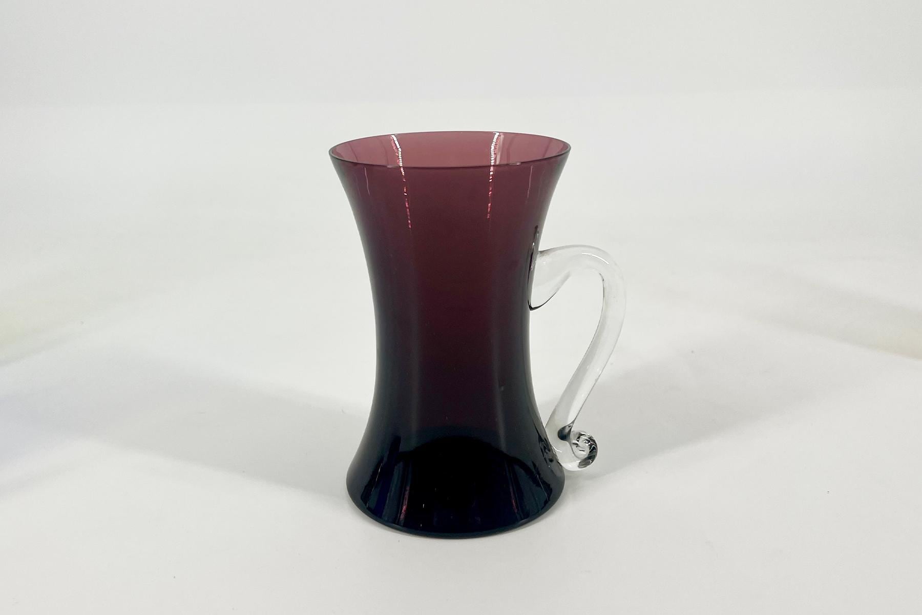 Glass pitcher with burgundy-red liquid and a clear curved handle on a white background.