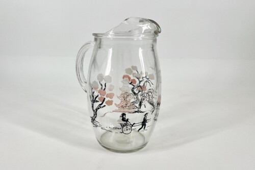 Clear glass pitcher with a lid and handle, featuring an Asian-style landscape illustration with trees, a pagoda, and small figures.