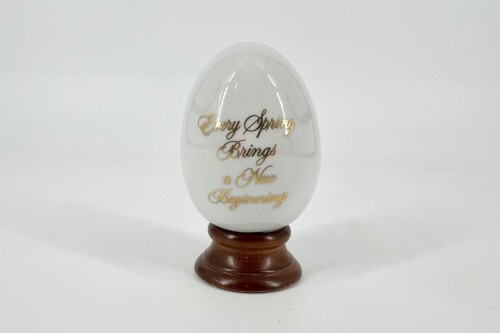 White egg-shaped decorative piece on a wooden stand with gold cursive text: 'Every Spring Brings a New Beginning'.