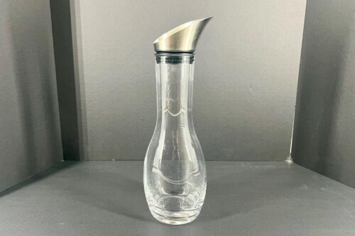 Clear glass water carafe with a stainless steel angled pour spout on top, set against a dark backdrop.