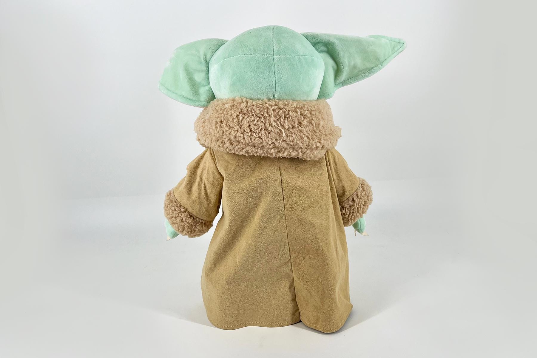 Back view of a Grogu plush toy wearing a tan robe, with green ears sticking out to the sides.