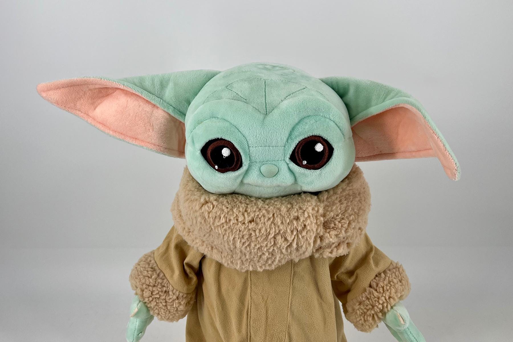 Close-up of a Grogu (Baby Yoda) plush toy with mint-green head, large brown eyes, and oversized pink-tipped ears in a tan robe.
