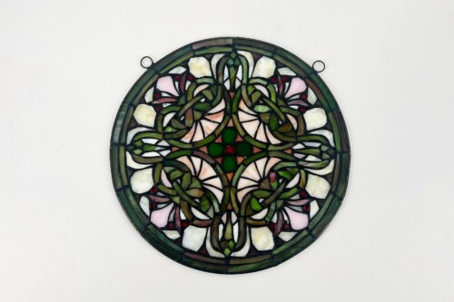 Round stained-glass panel with green vines and pale pink-white flowers, decorative floral design, hanging by metal loops at the rim.