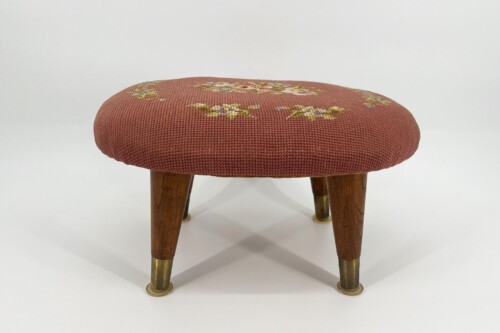 Round upholstered footstool with red fabric and floral embroidery, supported by wooden legs with brass caps.