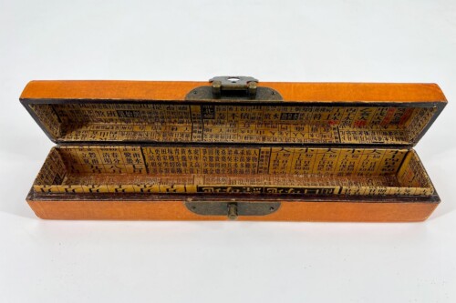 Open orange wooden box with a hinged lid, interior lined with yellowed pages of Chinese characters arranged in grid-like blocks, like a storage case for printing blocks or text tiles.
