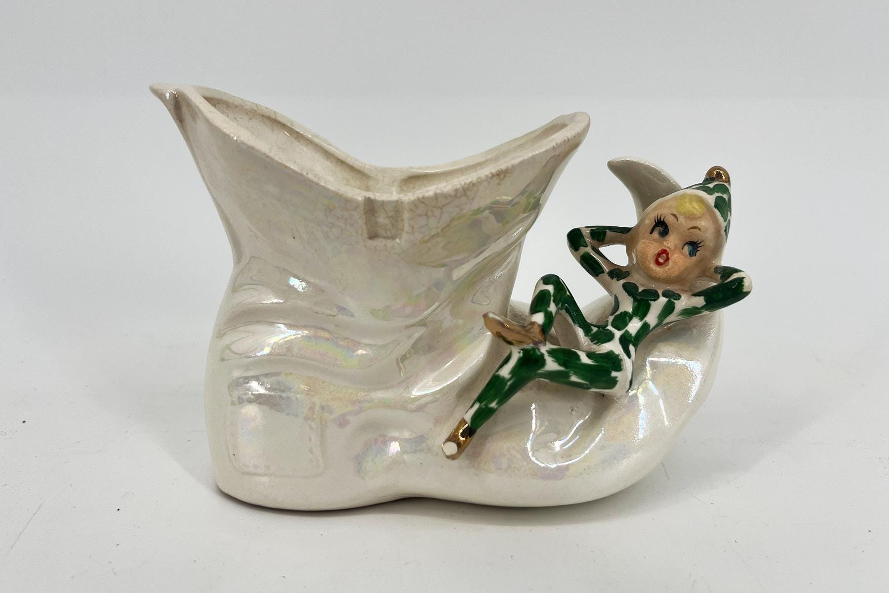 White glossy ceramic sculpture featuring a small figure in a green striped outfit clinging to a large curved, boat-like form.