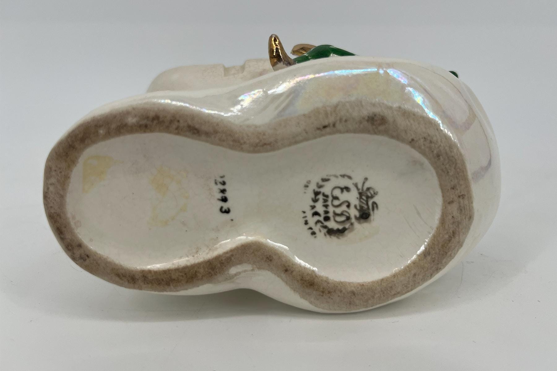 Bottom of a glossy ceramic figurine showing a curved hollow base, beige glaze, and maker's marks/stamps on the bottom edge with iridescent highlights nearby.