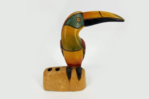 Wooden toucan carving perched on a square wooden stand with a long beak.