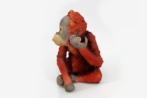 Clay monkey figurine with red-orange fur and gray face, sitting and turning to the left, one hand raised near its ear for listening