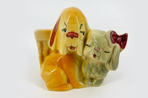 Ceramic figurine of a yellow dog leaning over a cream-colored cat wearing a red bow, both cuddling together.