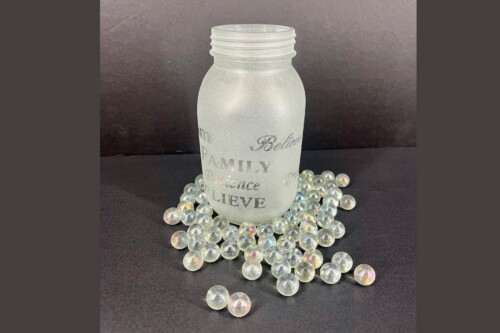 Frosted glass bottle with inspirational words like 'Family' and 'Believe' on a dark surface, surrounded by iridescent glass marbles.