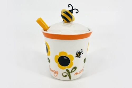Decorative ceramic cup with orange rim, yellow flowers and bee motifs, yellow spoon, and a lid topped by a bee ornament against a white background.