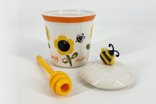 White ceramic cup with an orange rim and yellow flower pattern, bee motif, next to a white lid and yellow toy spurting from the foreground.