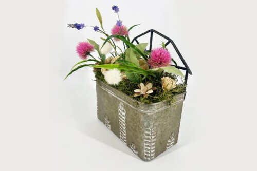 A vintage metal toolbox filled with a bright floral arrangement including pink thistles, white pom-poms, lavender sprigs, green leaves, and moss, with a black geometric handle.