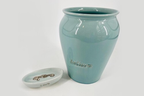 Turquoise ceramic vase with 'Believe' embossed, beside a small dish holding a silver bracelet on a white background.