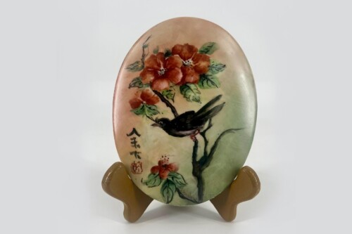Oval ceramic plaque with a black bird perched on a branch amid red flowers and green leaves; Chinese characters along the left edge.