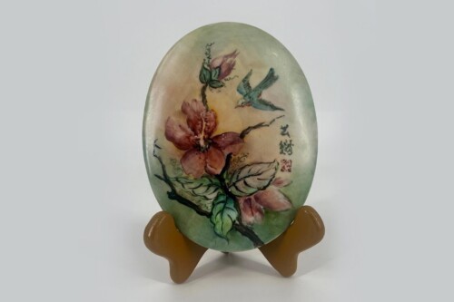 Oval decorative plate with pink flowers, green leaves, and a blue-green bird on a pale background, displayed on a brown stand.
