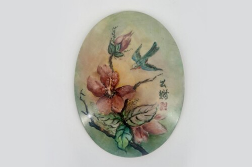 Decorative oval plate featuring a pastel painting of pink flowers, green leaves, and a blue-green hummingbird in soft background hues, with signature marks on the side.