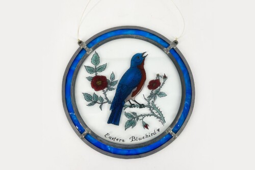 Circular stained-glass plaque showing an Eastern Bluebird perched on a flowering branch, blue rim surrounding the scene.