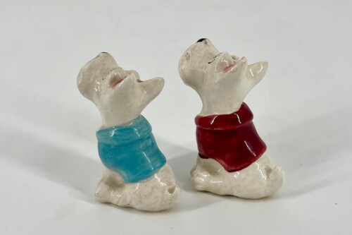 Two ceramic dog figurines with blue and red scarves looking upward at each other on a plain background