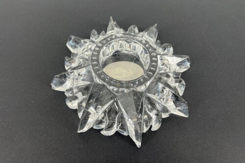 Clear crystal sunburst ornament with a central circular holder, resembling a starburst made of faceted prisms on a dark surface