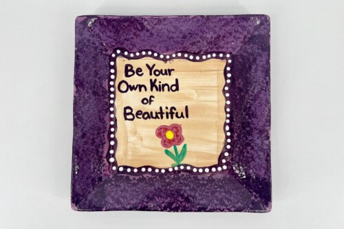 Purple decorative plaque with a beige center reading 'Be Your Own Kind of Beautiful' and a small red flower at the bottom.
