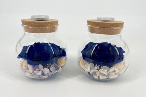 Two clear glass jars with cork stoppers, each containing blue artificial flowers and pale decorative petals.