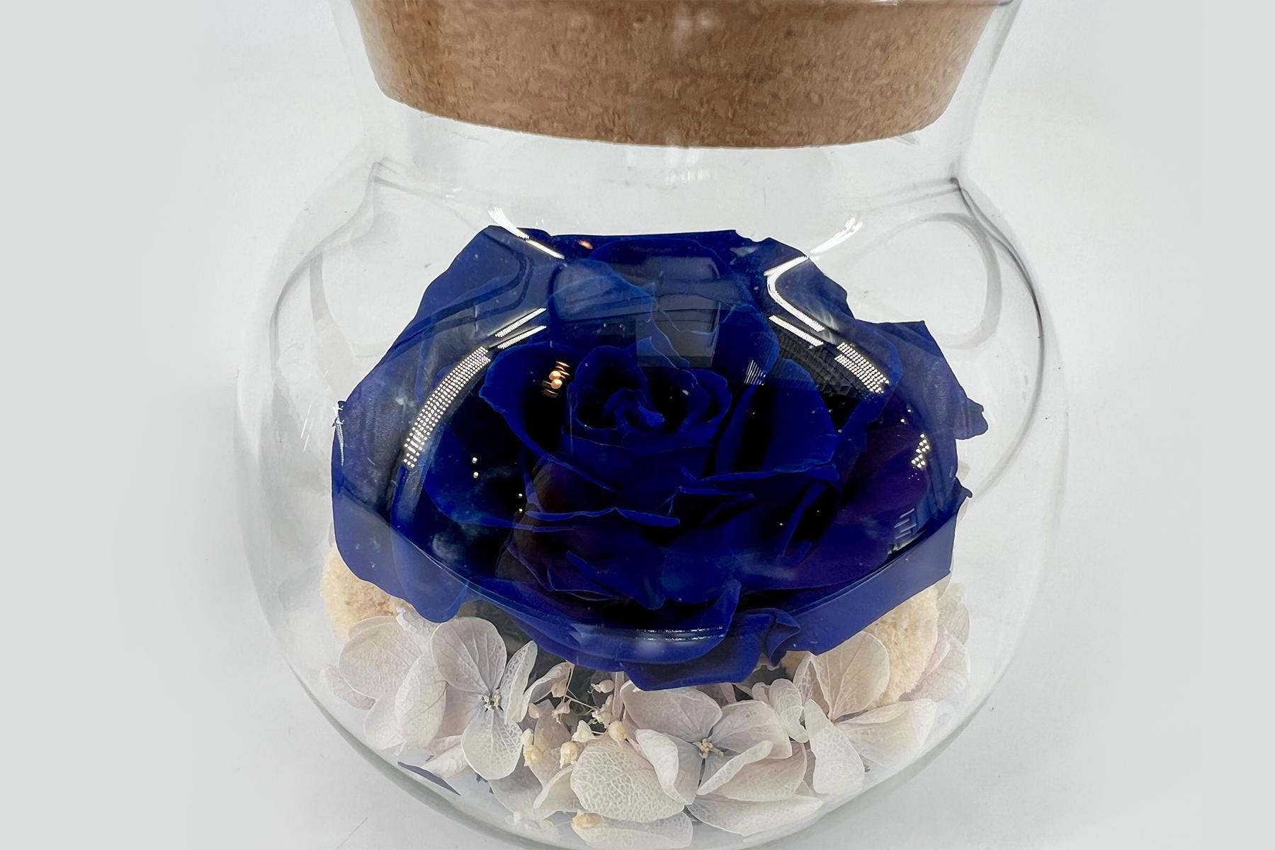 Clear glass jar with a deep blue rose encased inside, and pale dried flowers at the bottom beneath the rose.
