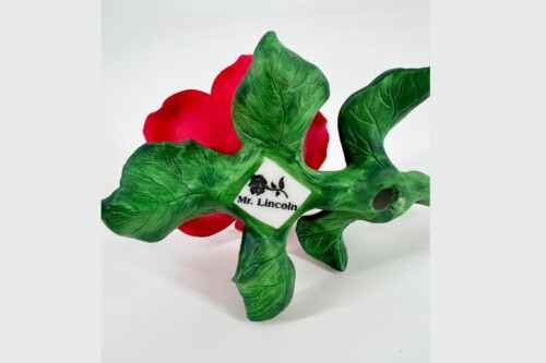 Decorative masquerade mask with green leaf-shaped pieces and a red background, labeled 'Mr. Lincoln' on a white tag.