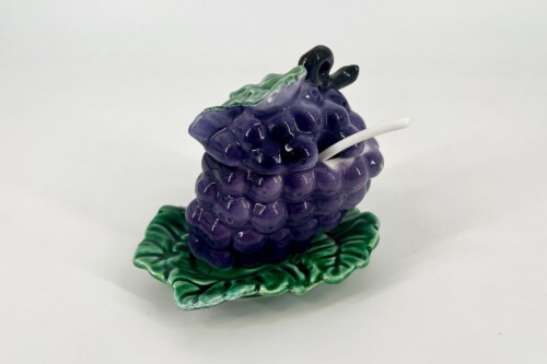 Glossy ceramic sculpture of a purple grape cluster resting on green leaves, with a white stem-like piece protruding from the cluster.
