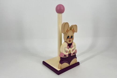 Wooden bunny toy wearing pink and purple clothes stands beside a tall wooden post with a pink ball on top.