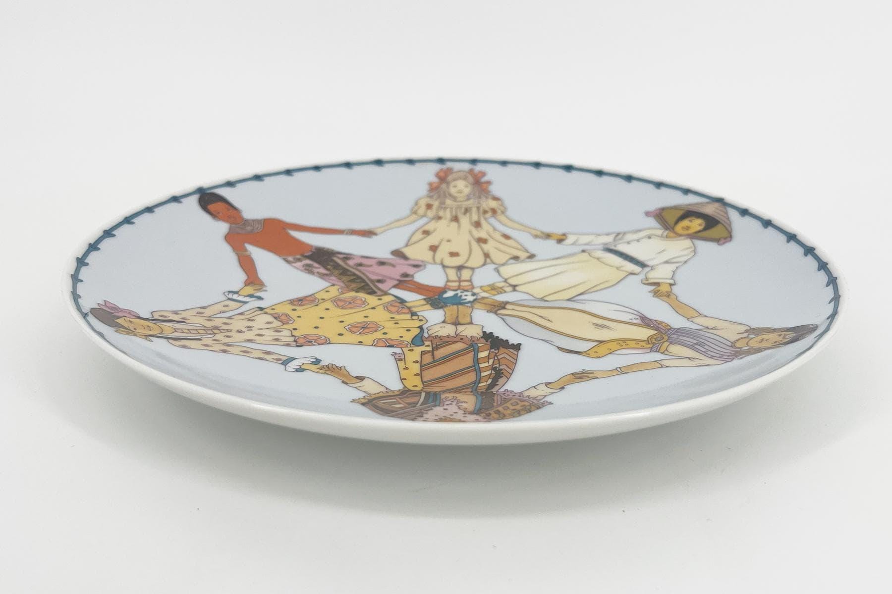 Decorative porcelain plate with a circular illustration of people in colorful traditional dress dancing in a ring, edged with a stitched blue border.