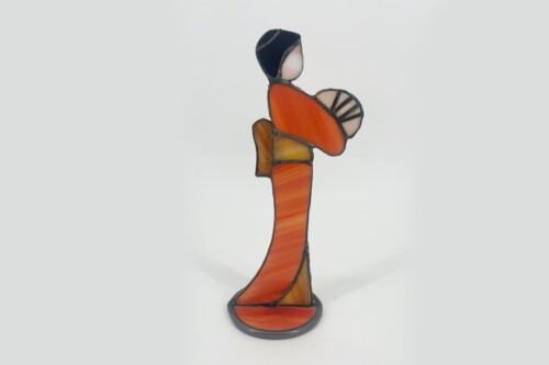 Abstract stained-glass sculpture of a tall figure in an orange dress, with dark hair, lifting one arm raised above the head and standing on a round base.