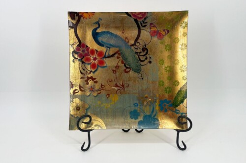 Decorative square plate with a blue peacock and colorful flowers on a gold background, displayed on a black metal stand.