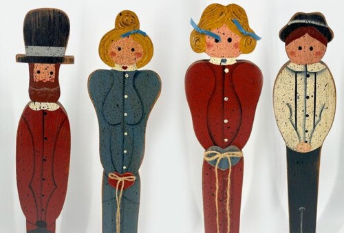 Four wooden folk-art figures standing in a row: a man with a top hat, two women in blue and red dresses, and a person in a light jacket with a dark cap.