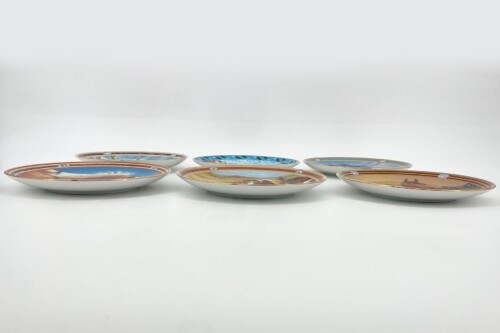 Six decorative porcelain plates with colorful rim designs arranged on a white surface, photographed in a studio setting.