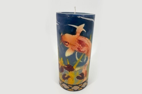 Tall decorative candle with an orange koi fish design swimming among yellow flowers on a dark blue background.