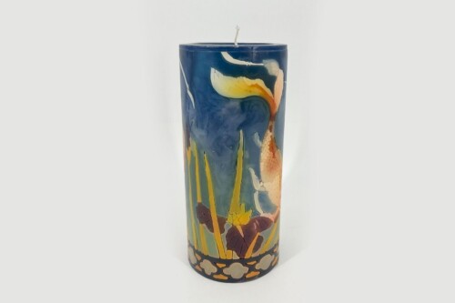 Tall cylindrical decorative candle with a blue landscape motif, featuring yellow flowers and tall grasses at the base and a visible wick at the top