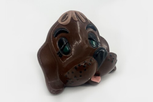 Brown ceramic dog figurine resting on its paws with a sad, droopy-eyed expression and pink tongue showing.