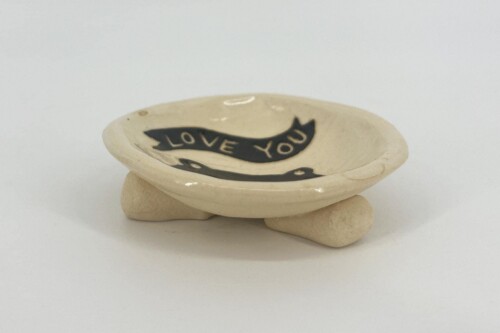 Beige ceramic bowl resting on small stones with a black ribbon inside that reads “LOVE YOU.”