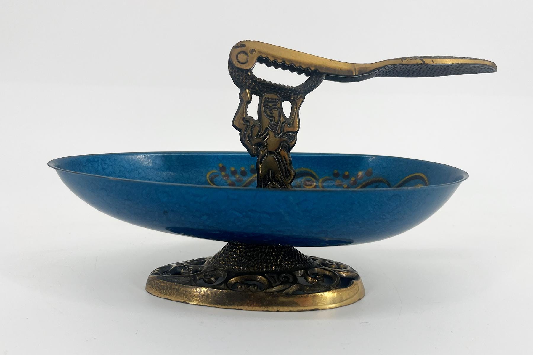 Ornate metal pedestal supporting a cobalt blue ceramic bowl; a sculpted figure rises from the center, holding a jagged, lever‑like bar across the top.