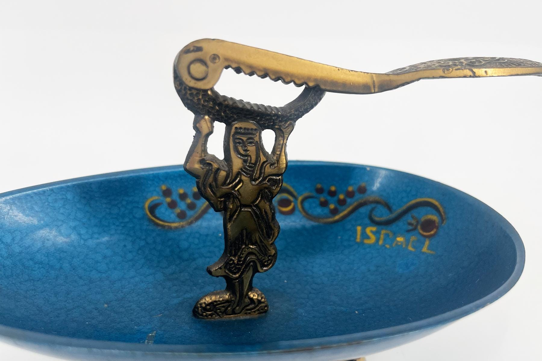 Bronze-toned metal figurine of a man raising a crocodile-head sculpture over his head, placed in a blue bowl with decorative patterns and 'Israel' inscription visible in the background.