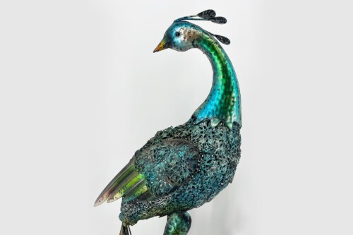 Colorful metal sculpture of a peacock with blue-green iridescent finishing and intricate lace-like feathers against a pale background.